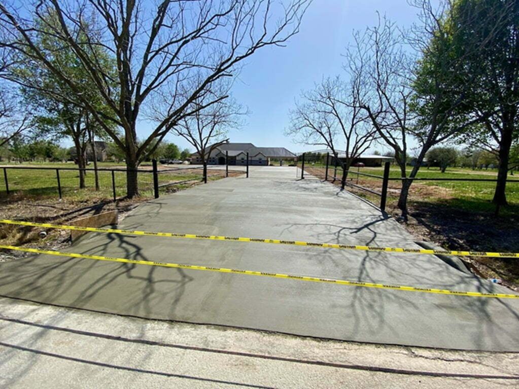 Freshly poured concrete driveway blocked by yellow caution tape, leading to a house in a rural setting.
