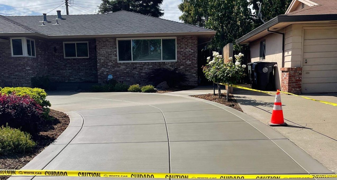 Freshly poured concrete driveway in front of a brick house, bordered by plants, with caution tape.