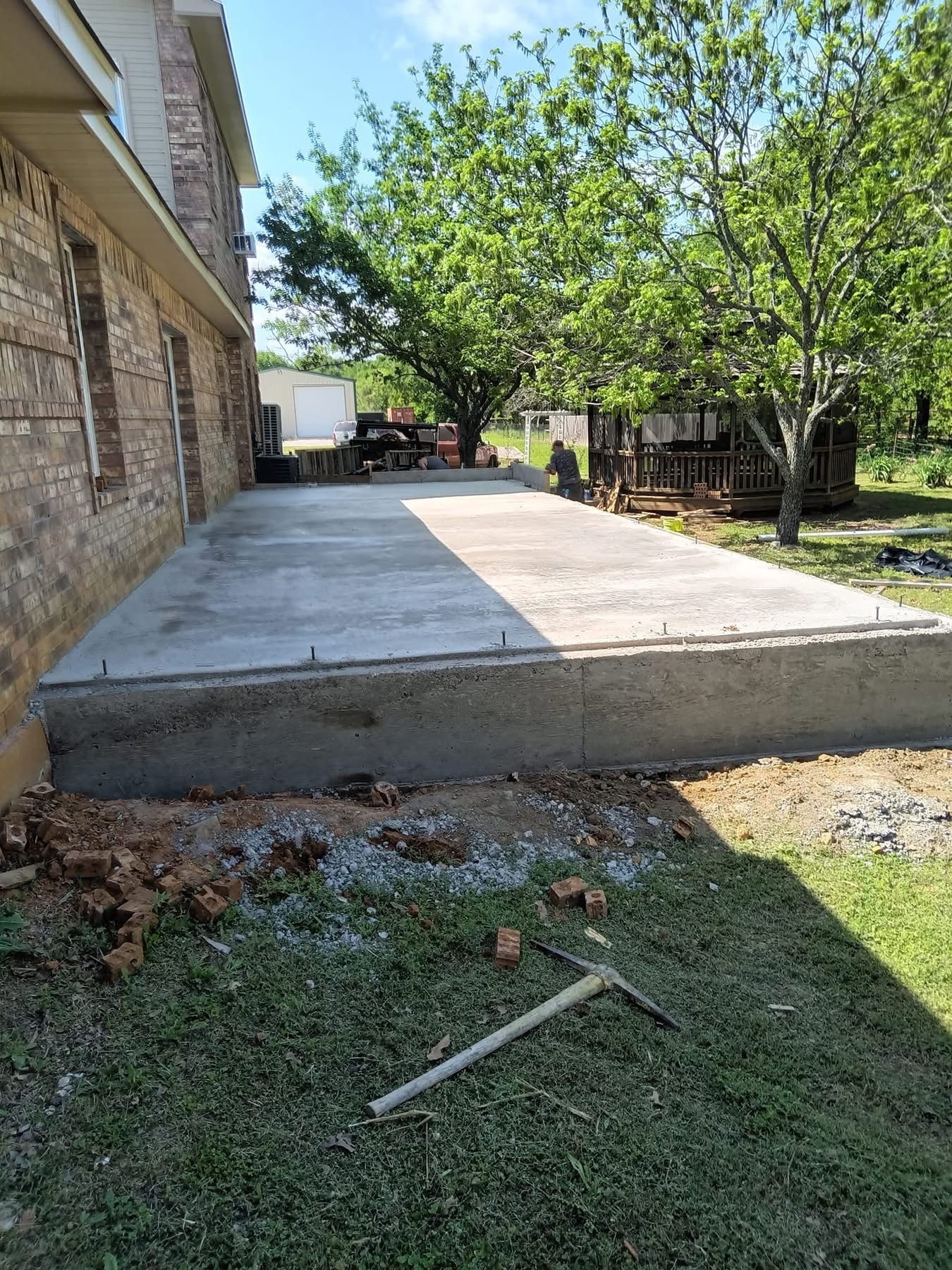 Concrete patio next to a brick building. Lawn with pickaxe in the foreground. Trees and a truck are in the background.