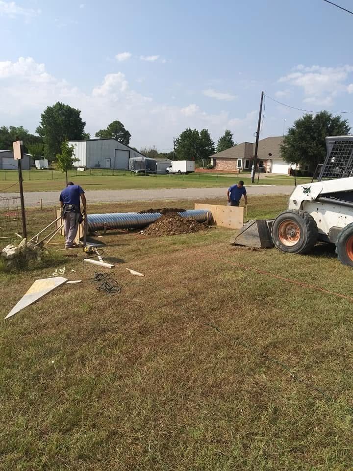 Two workers near an excavation site, a bobcat loader, and a pipe on grass under a cloudy sky.
