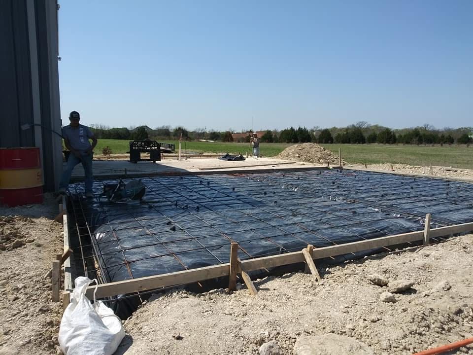 Construction site: Workers near a concrete foundation with black mesh and wooden forms, under a blue sky.