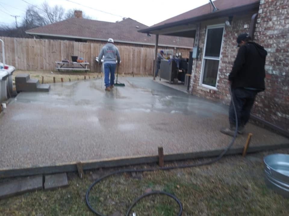 Men working on a wet concrete patio near a house. One is sweeping, another is standing.