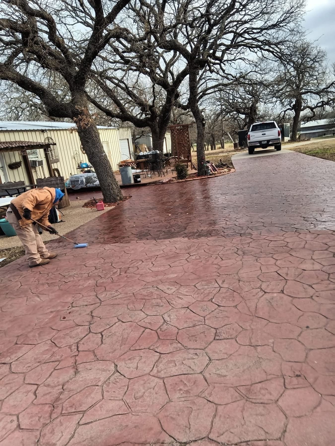 Person cleaning reddish, textured driveway near house and trees; truck in the distance.