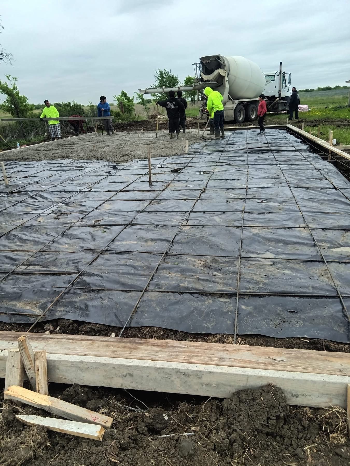Construction workers pouring concrete for a foundation, using a cement truck. Rebar and black plastic sheeting visible.
