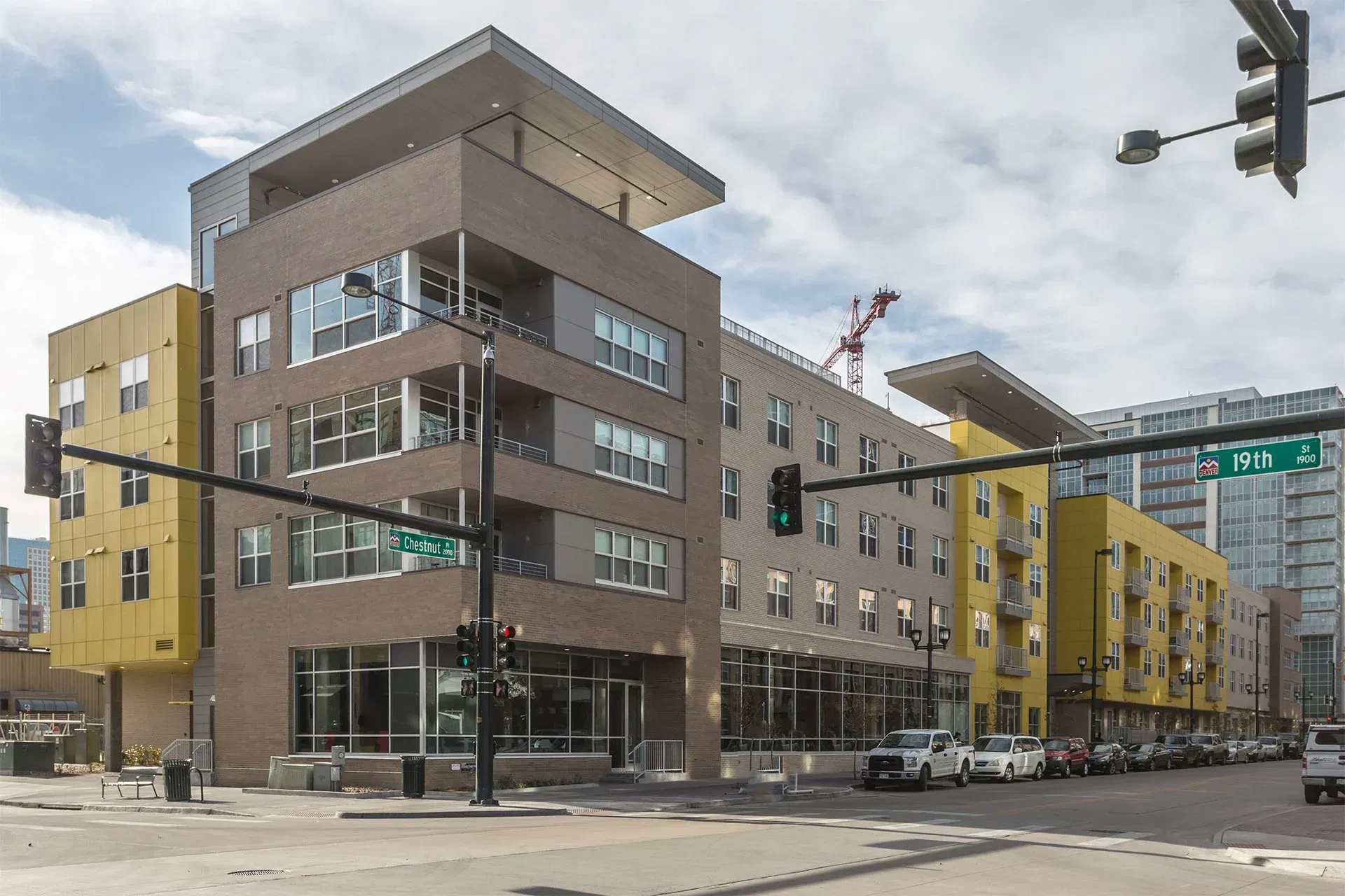 A modern, multi-story apartment building with yellow and gray facades on a sunny city street corner with traffic signals.