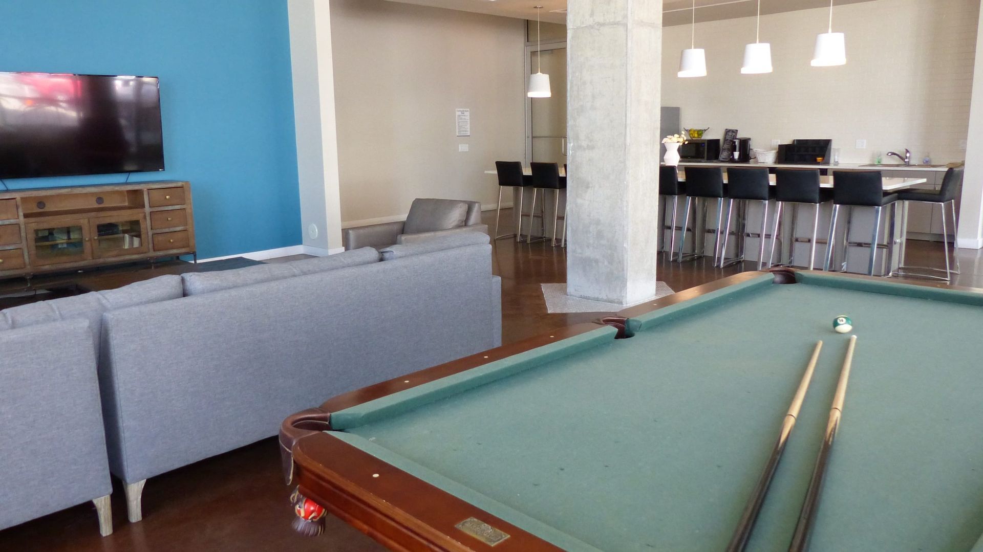 A lounge with a pool table in the foreground, a grey sofa, a television on a wooden stand, and a bar with stools behind.