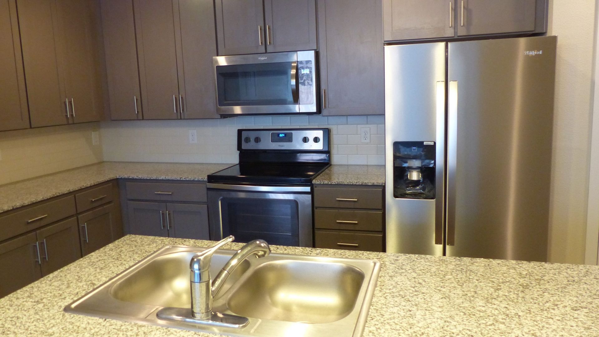 A modern kitchen with grey cabinets, stainless steel appliances, speckled granite countertops, and a double basin sink.