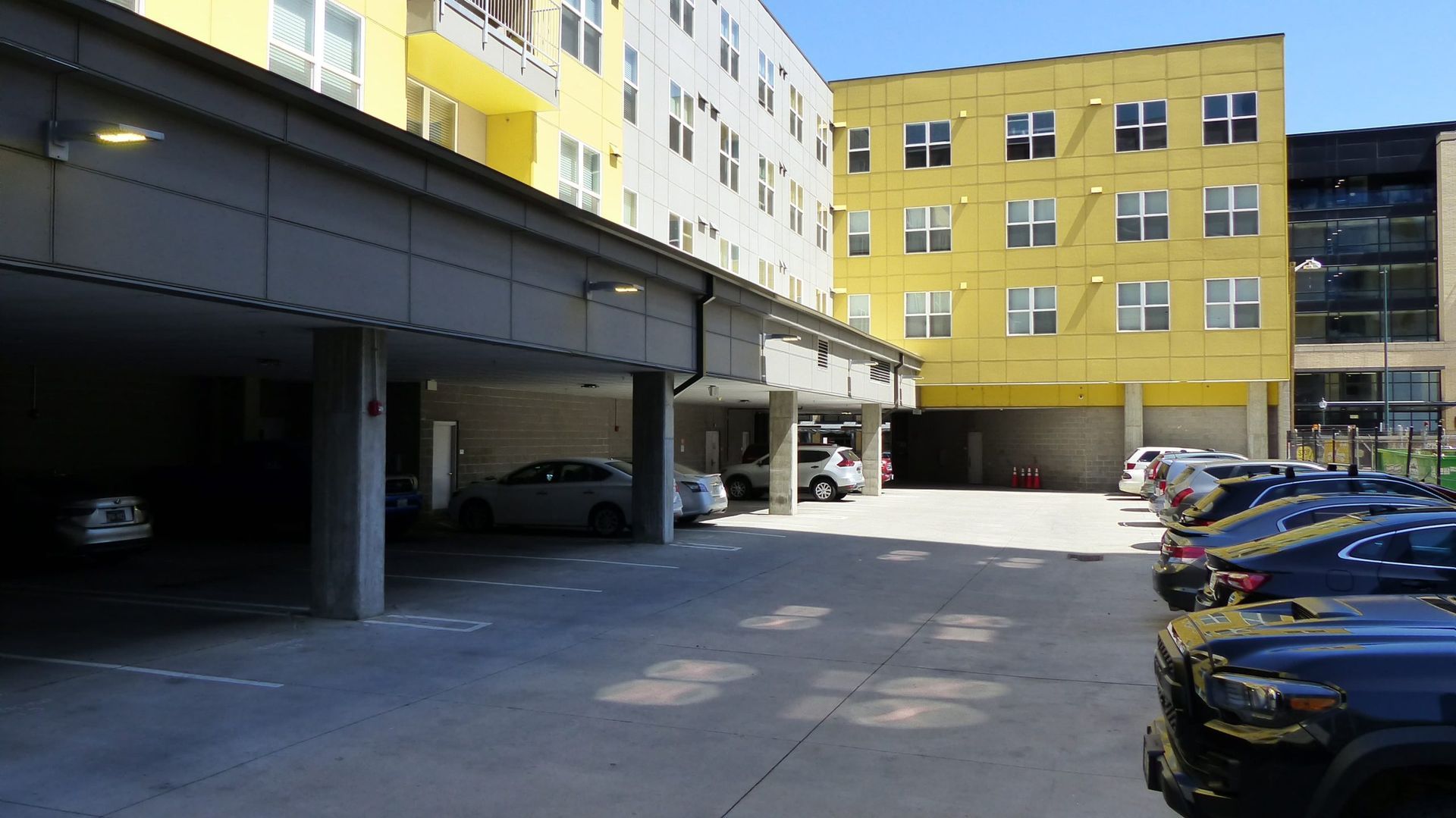 An outdoor parking lot beneath an apartment building with yellow siding, featuring cars parked in shaded and open spaces.