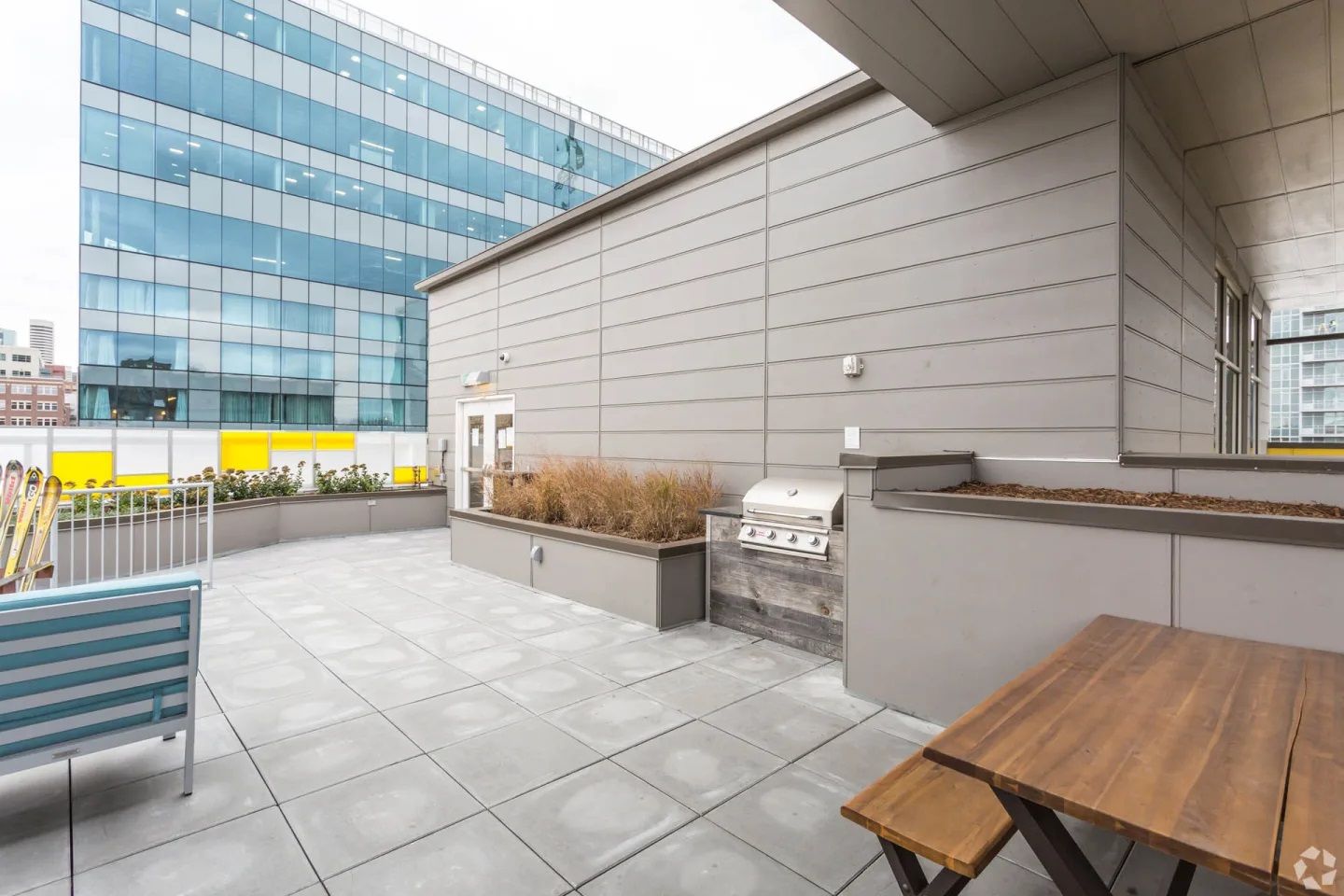A rooftop patio featuring a wooden picnic table, a built-in grill, planters with dried grass, and a view of a city building.