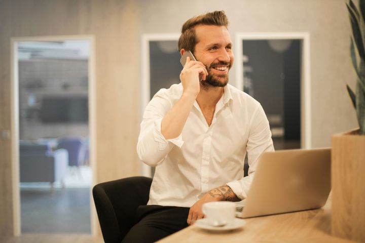 A person in a white button-down shirt talks on a phone while smiling, sitting at a wooden desk with a laptop and coffee.