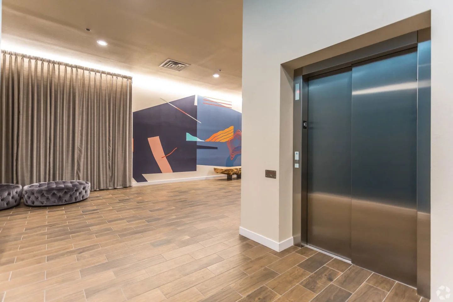 A modern lobby featuring an elevator, wood-look tiled floors, full-length curtains, and a colorful abstract wall mural.