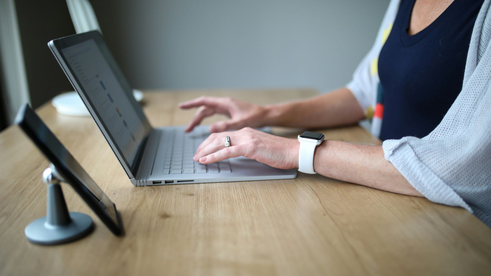 A person typing on a laptop at a wooden desk with a smartphone in a stand beside them.