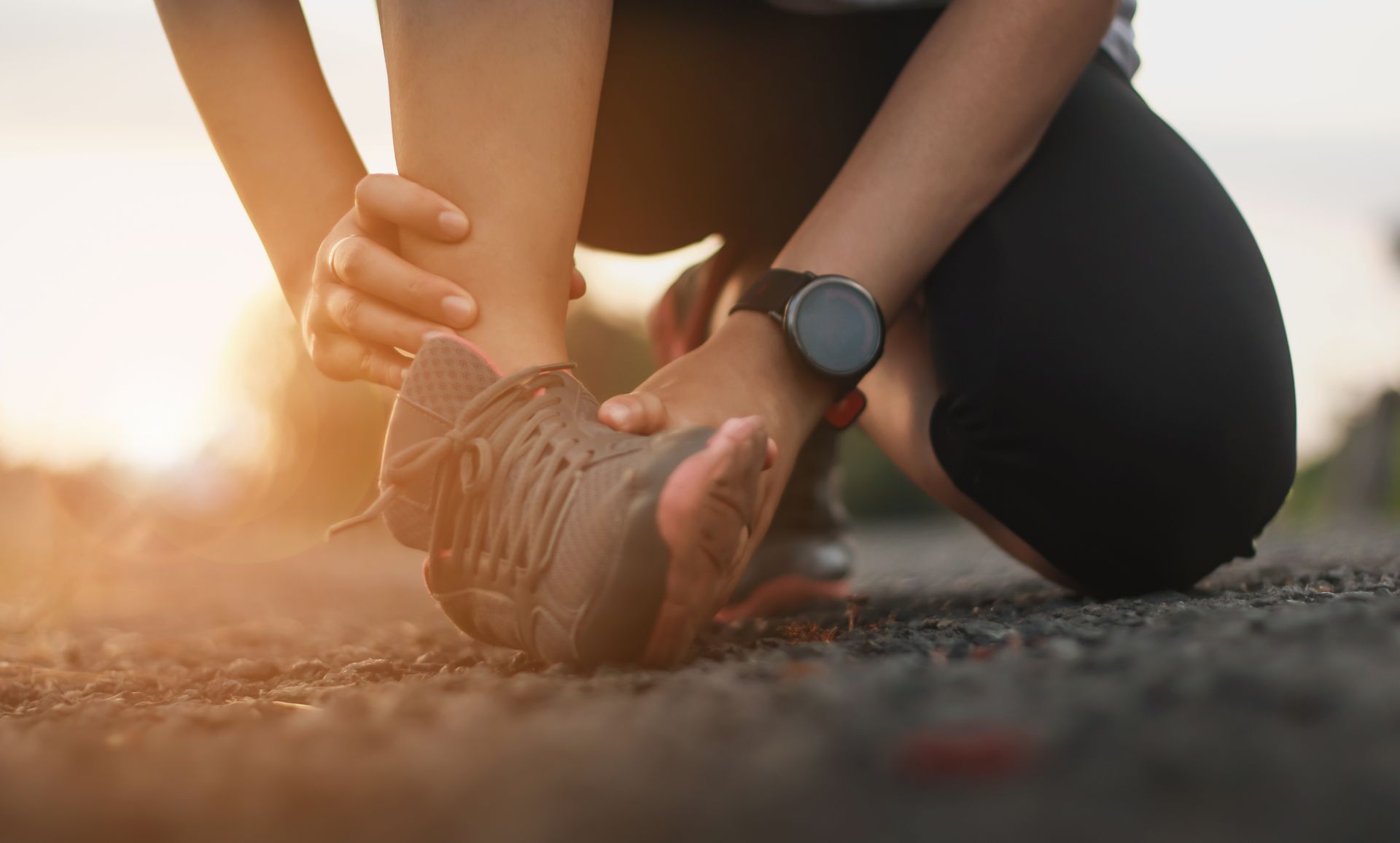 Person kneeling, holding ankle in pain after exercise. Sunset in background.
