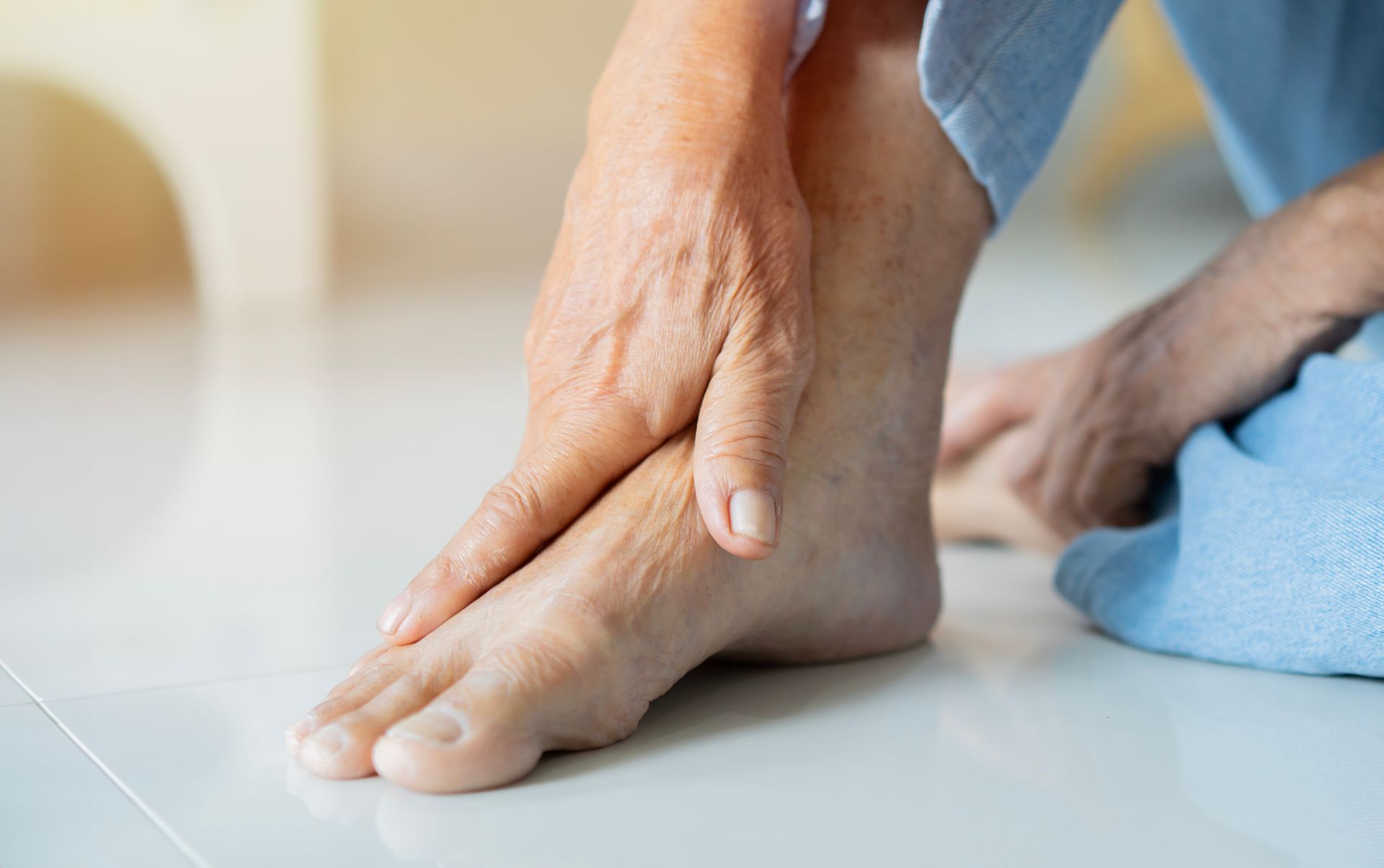 Person holding their foot and ankle, likely in pain, indoors, with a tiled floor.