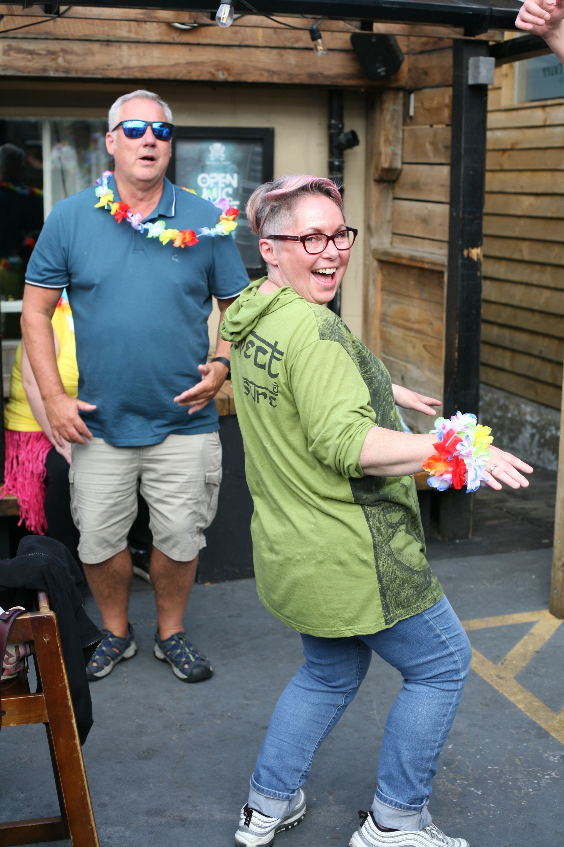A woman in a green shirt is dancing in front of a man wearing sunglasses.
