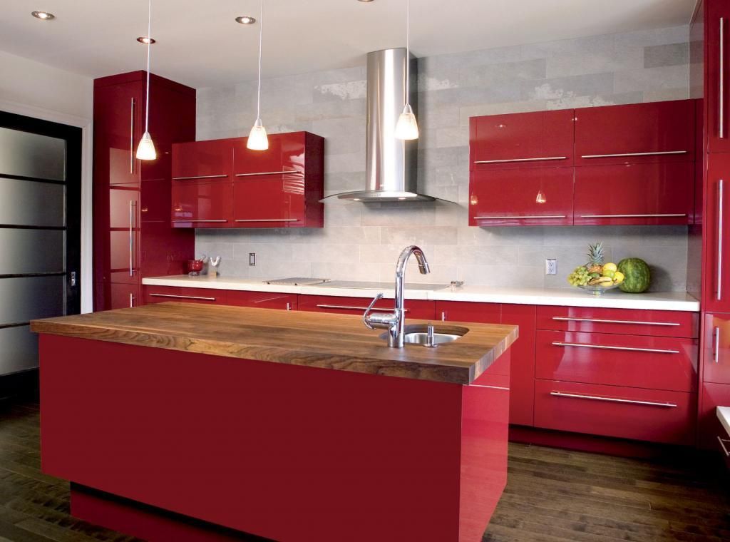 Cocina moderna con gabinetes de color rojo brillante, isla de madera, campana de acero inoxidable y luces colgantes.