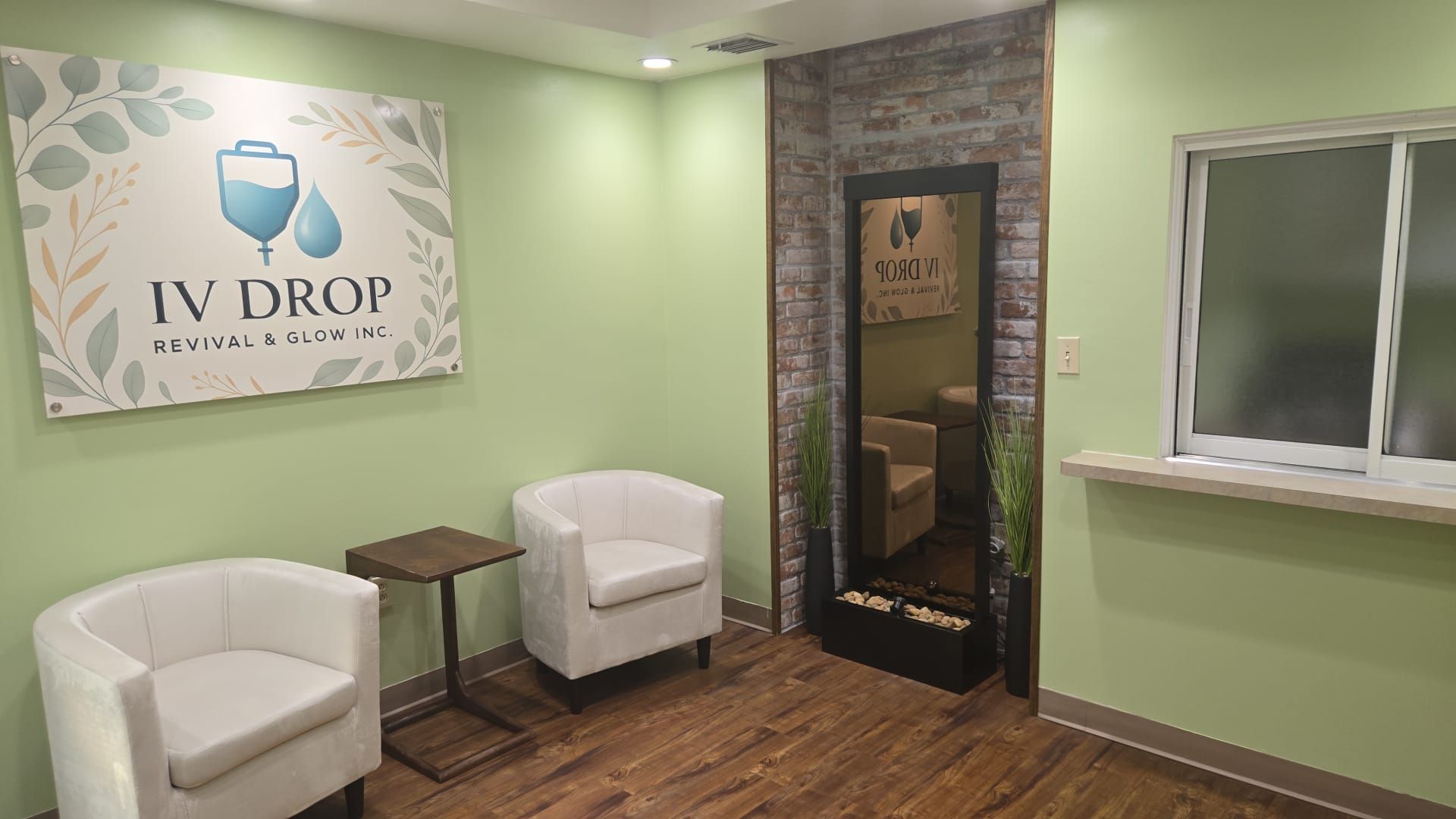 A waiting room with white chairs, a table, a mirror, and an IV Drop sign. Green walls and wood floor.