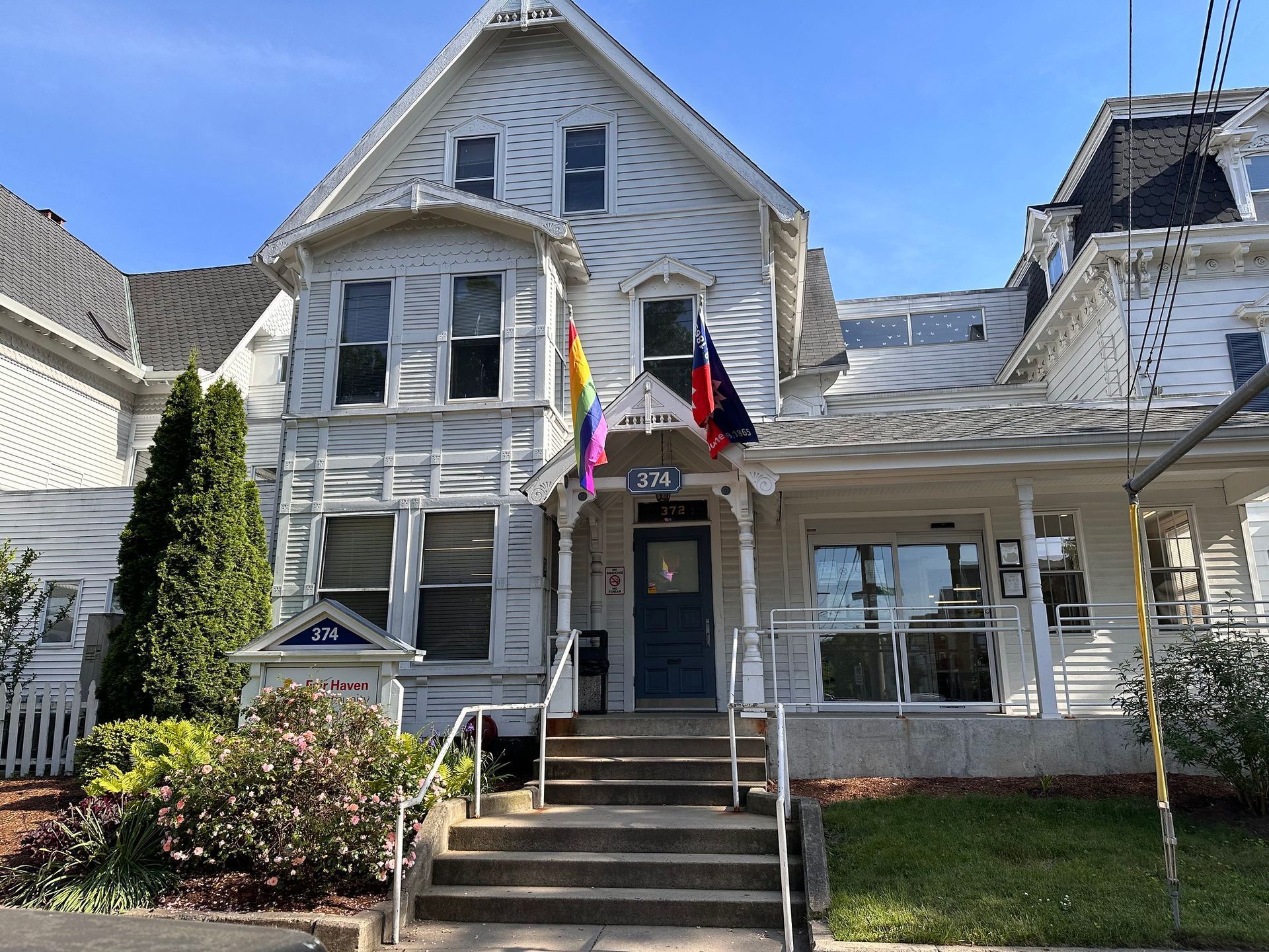 A white, multi-story Victorian-style building with a front porch, stairs, a Pride flag, and a US flag near the entrance.