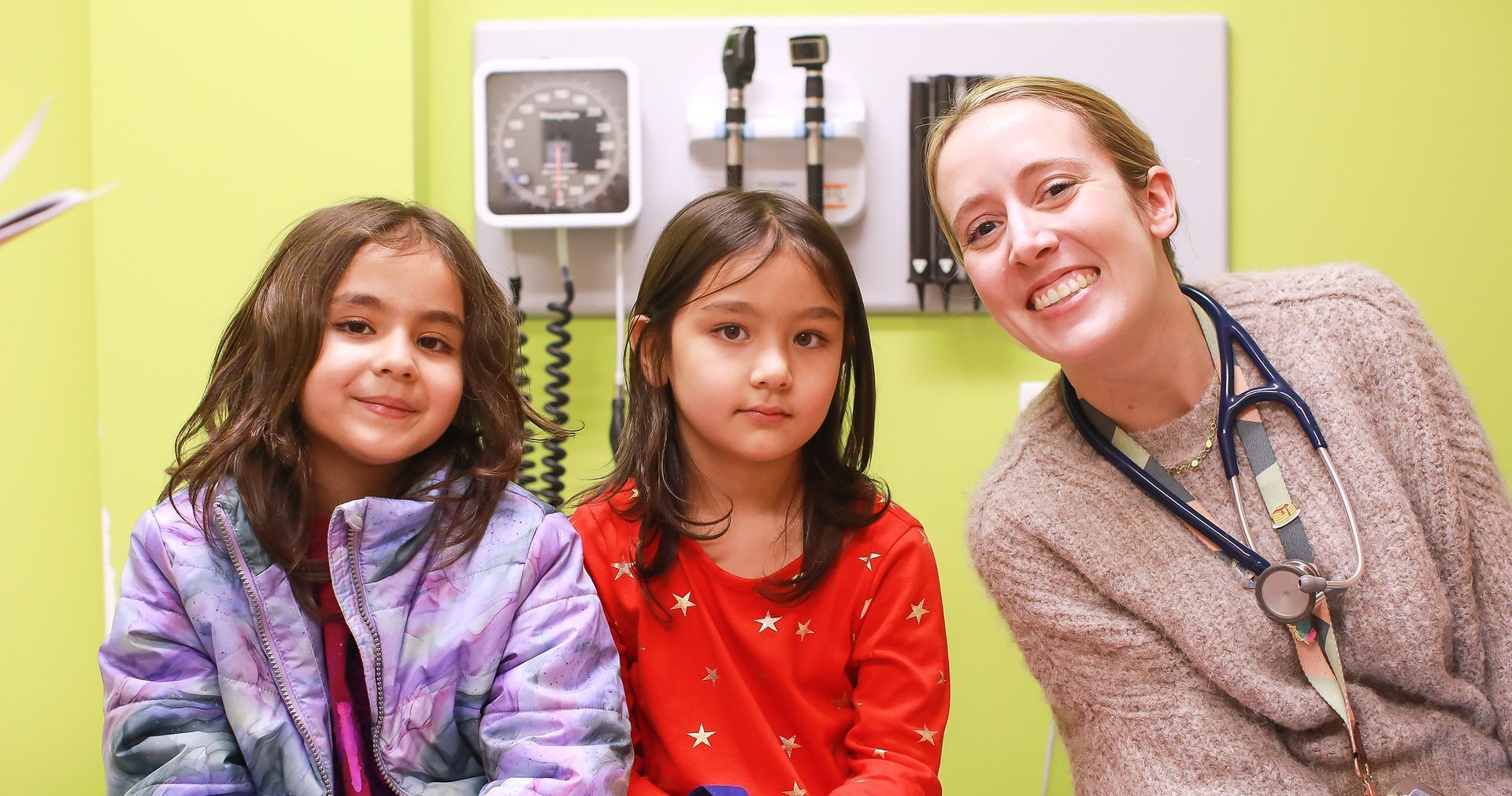 A provider wearing a stethoscope smiles with two young children in a lime-green clinical exam room.