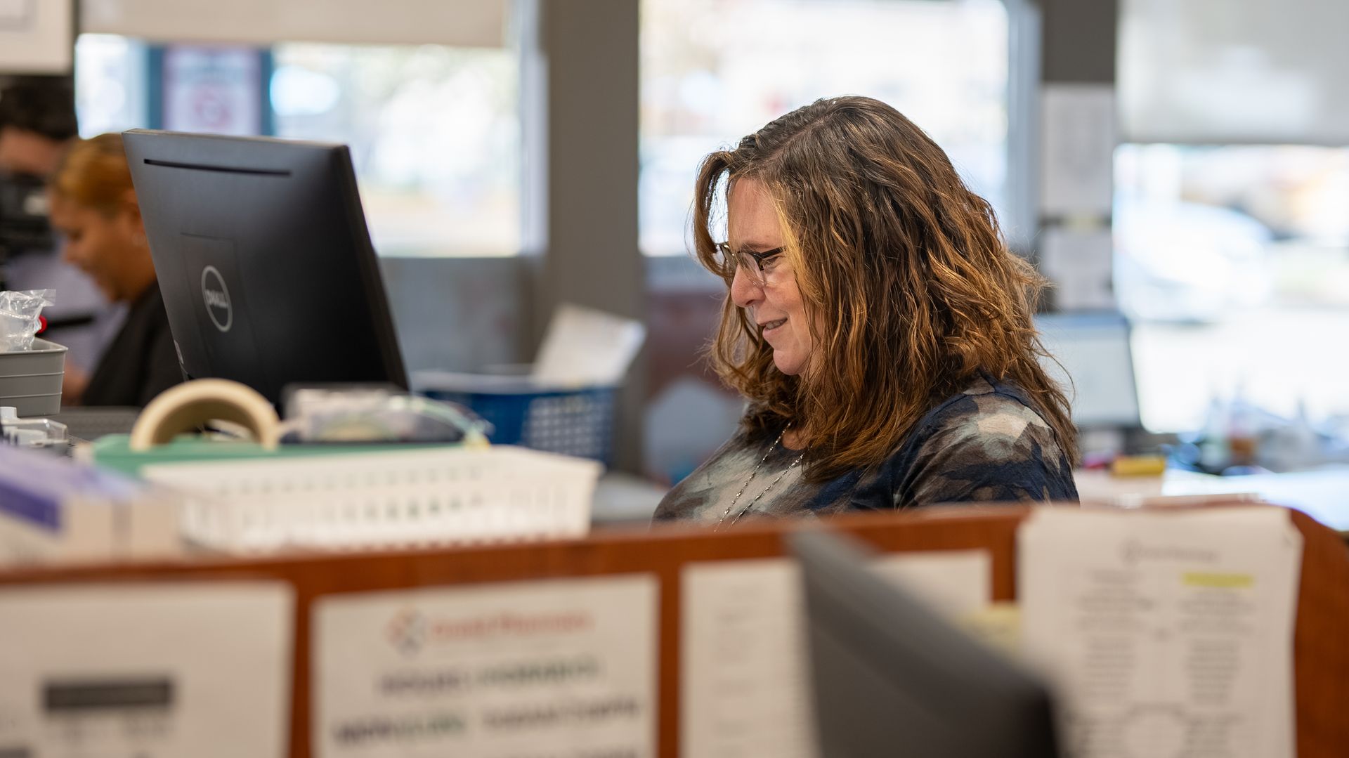 A person with long wavy hair, wearing glasses, works at a computer in a busy office setting.
