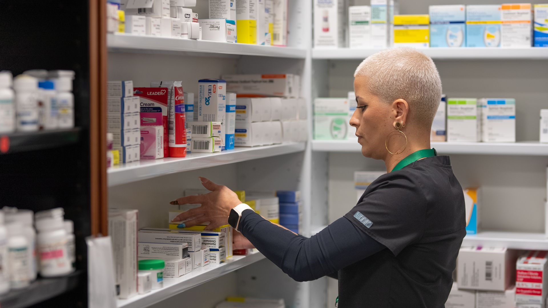 A pharmacy technician in black scrubs organizes medication boxes on a shelf in a brightly lit pharmacy.