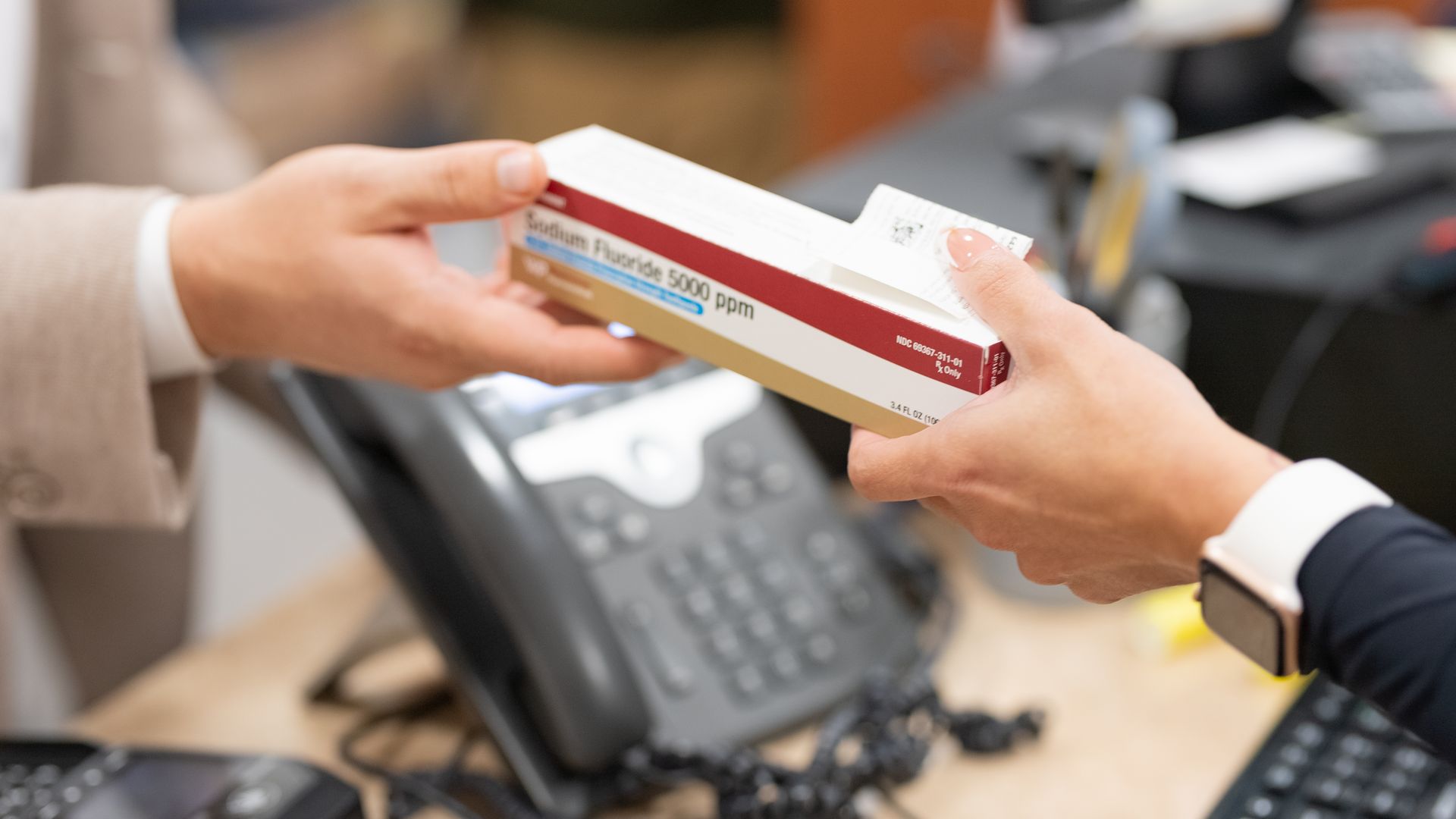 A person hands a box of prescription toothpaste to another person in an office setting.