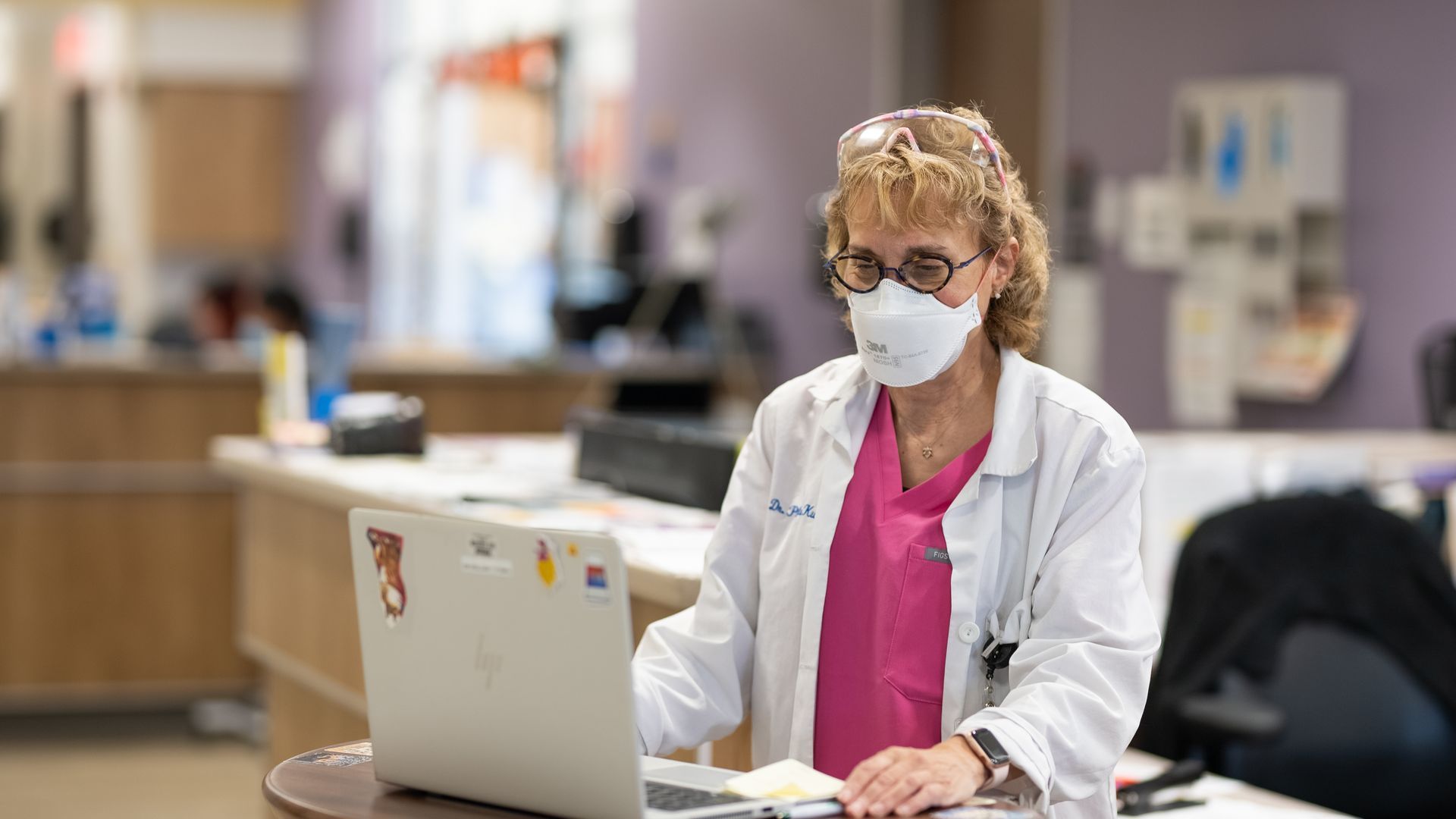 A healthcare worker in a white coat, pink scrubs, and face mask works at a laptop in a medical office setting.