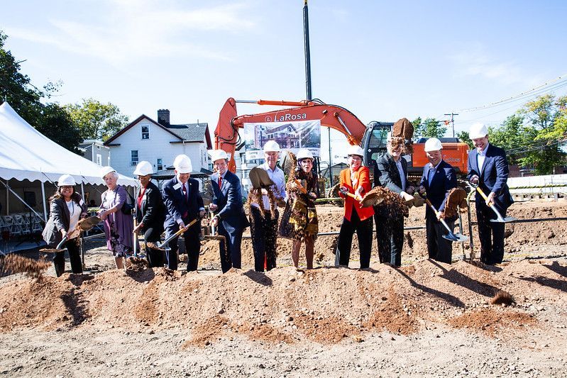 A group of people in hard hats participate in a ceremonial groundbreaking with shovels in front of an orange excavator.