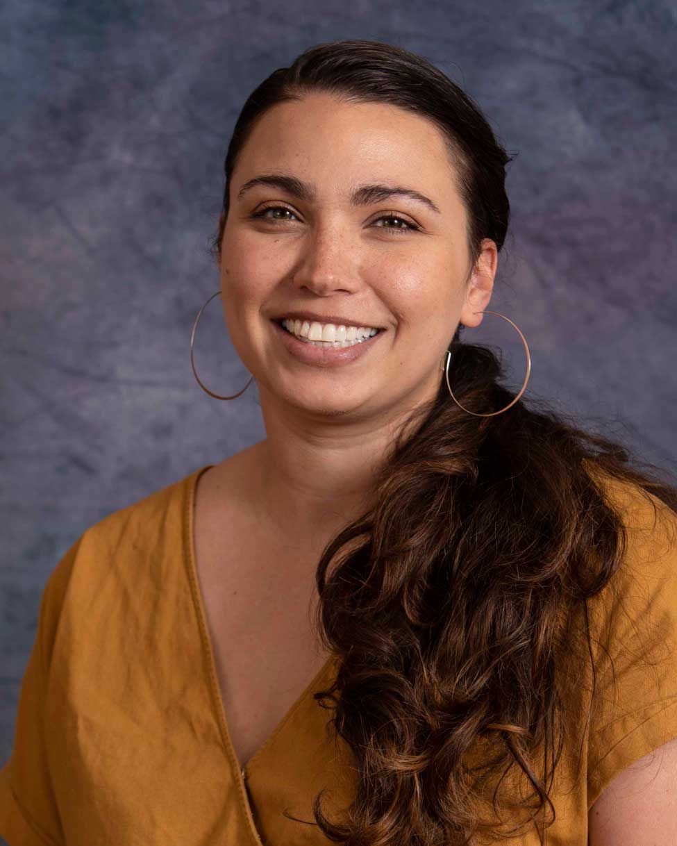A smiling person with long, dark wavy hair wears large gold hoop earrings and a yellow top against a mottled gray backdrop.