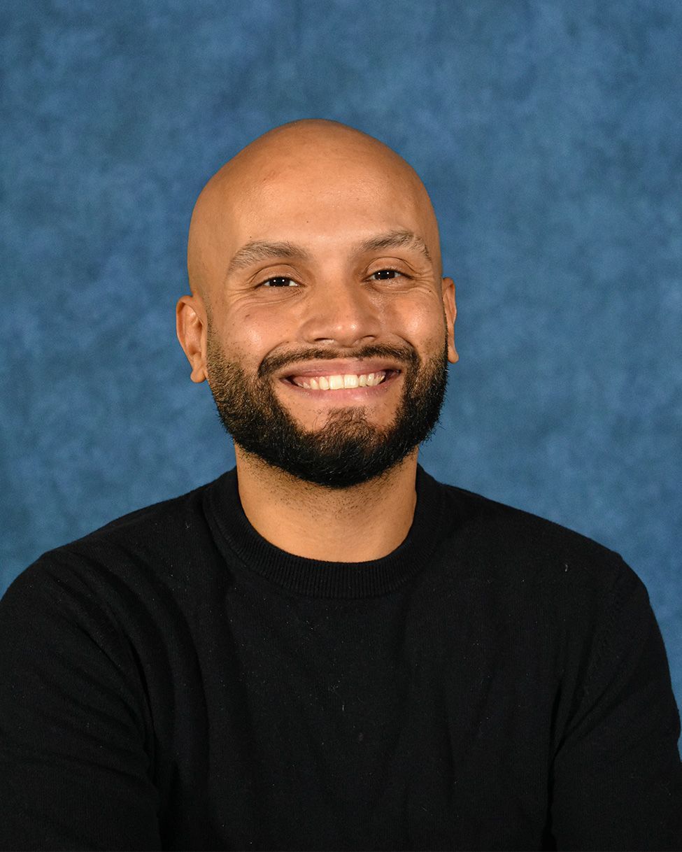 A smiling person with a short beard wears a black crew-neck sweater against a mottled blue background.