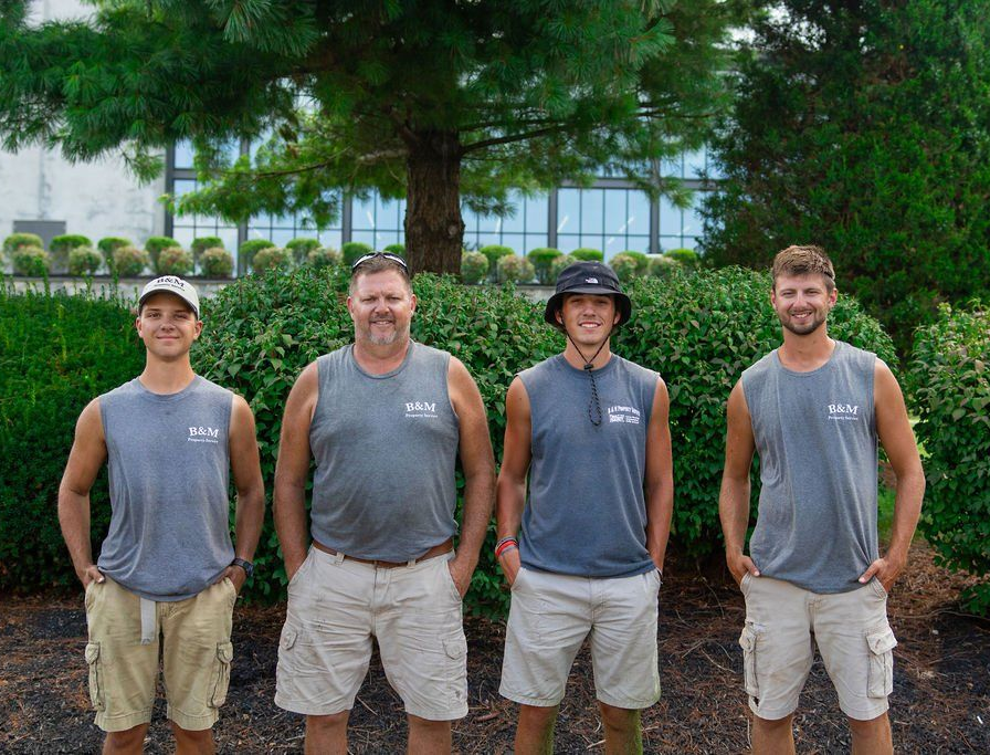 Four men wearing gray sleeveless shirts and khaki shorts stand in front of greenery.