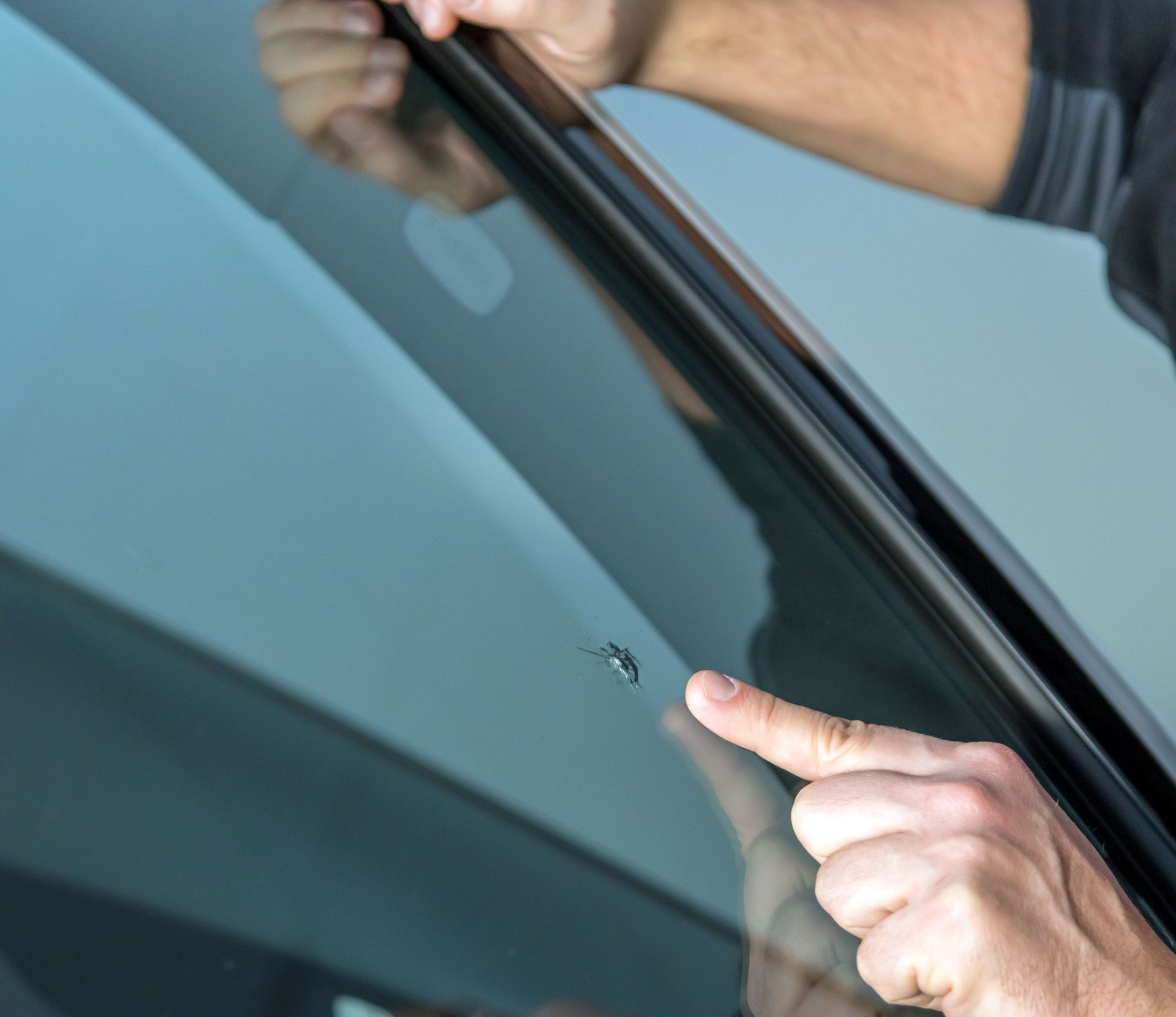 Person points at a small chip on a car windshield, highlighting the damage.