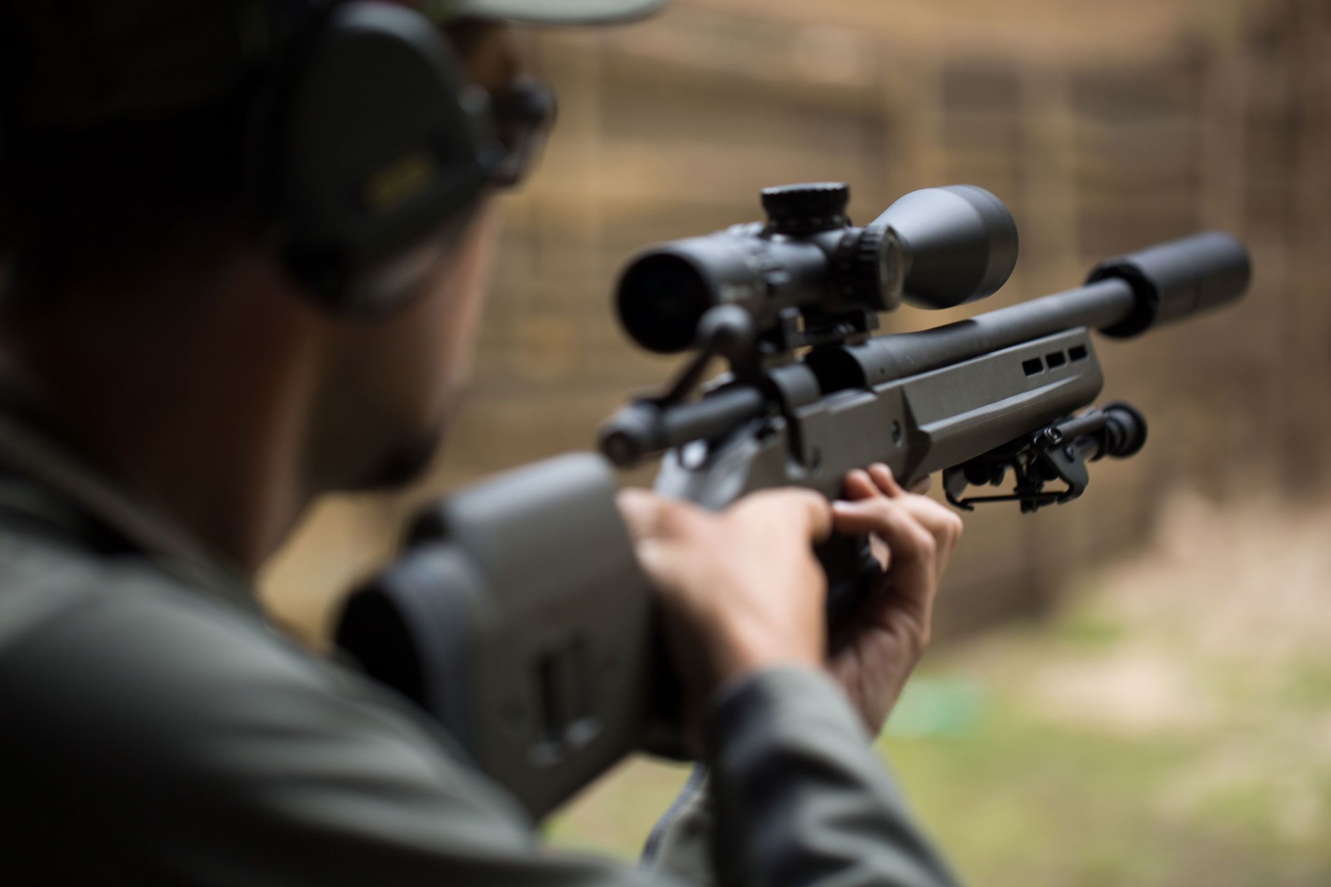 Person aiming a rifle with a scope at a shooting range, wearing ear protection.