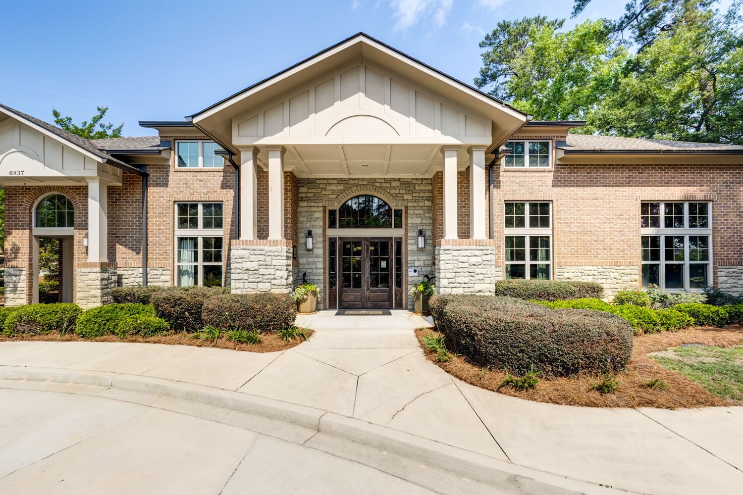 Brick building with a stone entrance, double doors, and windows, with bushes and a walkway at Arcadia's Edge in Columbia, SC.