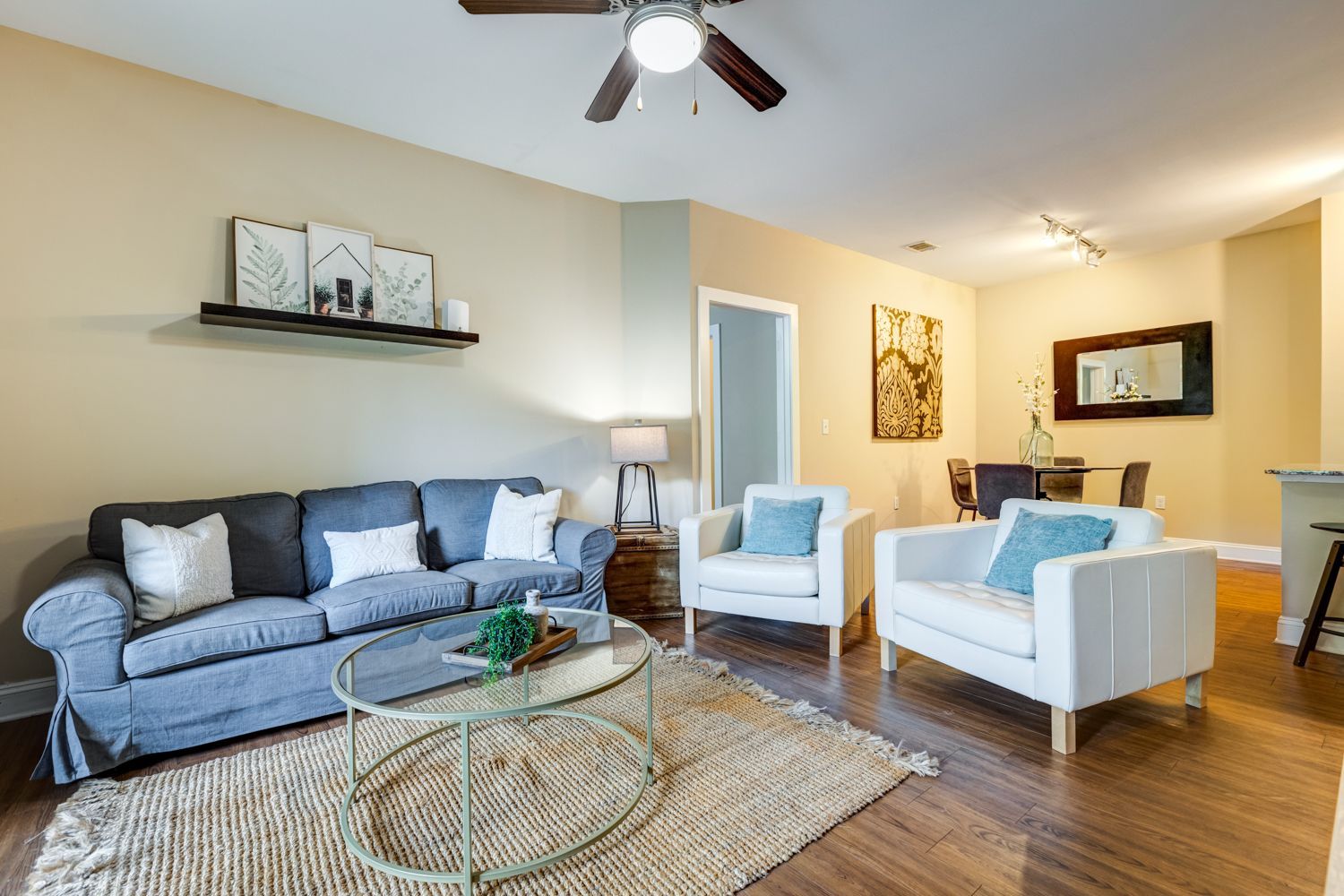 Living room with gray couch, white chairs, coffee table, rug, and dining area in the background at Arcadia's Edge in Columbia, SC.