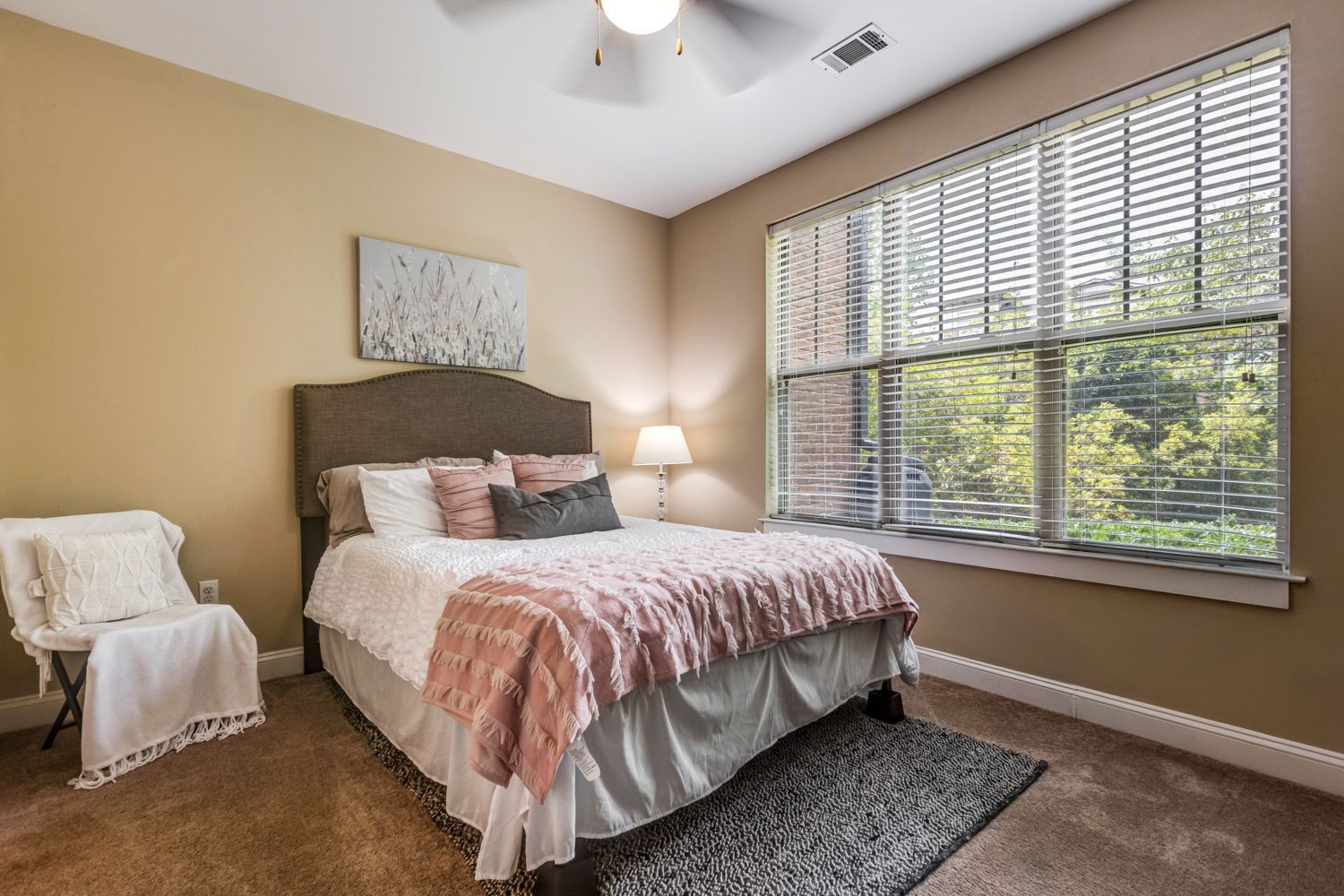 Bedroom with bed, chair, window, and neutral walls; brown carpet at Arcadia's Edge in Columbia, SC.
