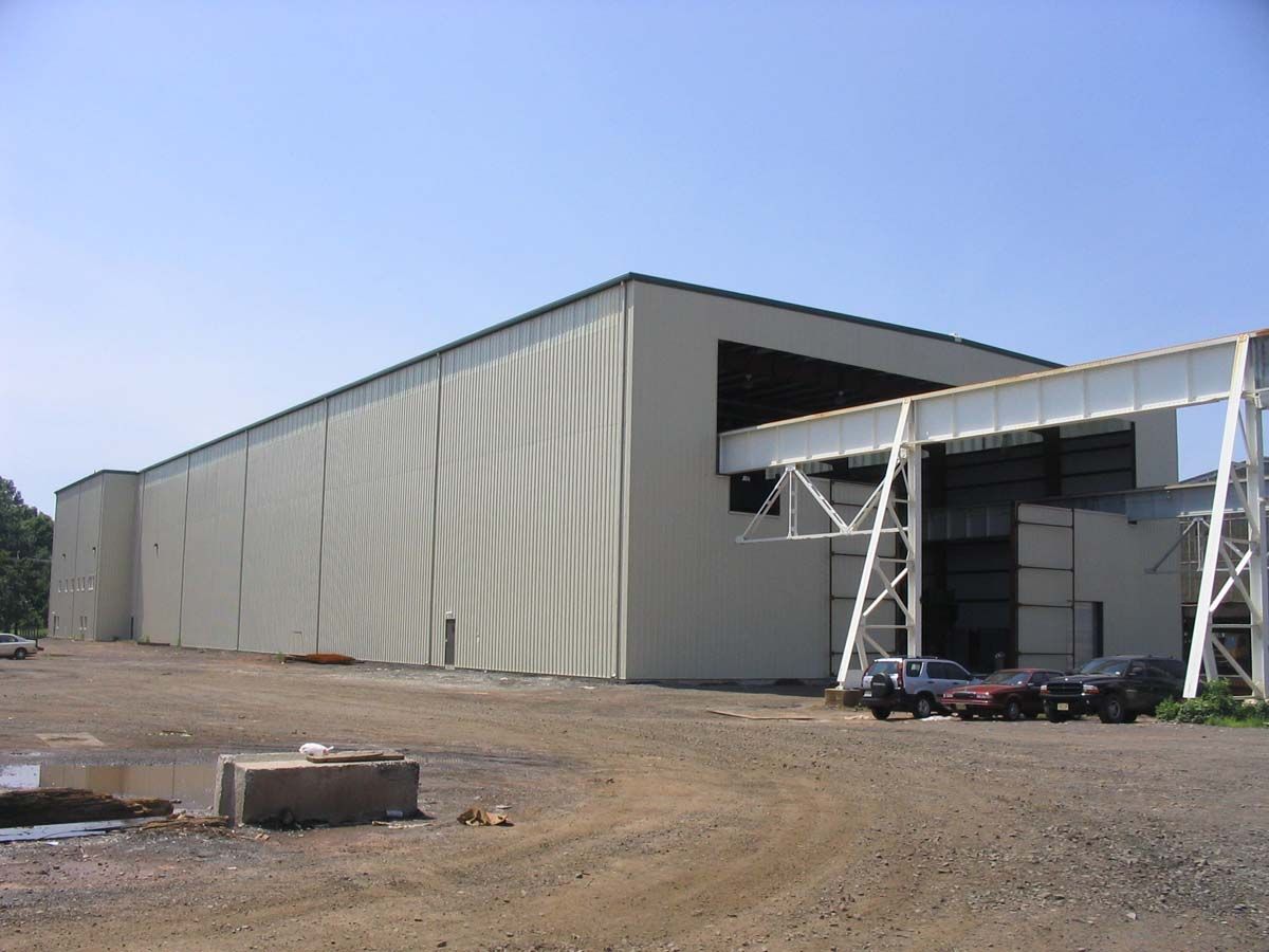 A large, beige, metal-sided industrial warehouse building with an exterior overhead crane structure in a gravel lot.