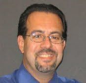 A person with a goatee and glasses wearing a blue shirt, smiling against a plain gray background.