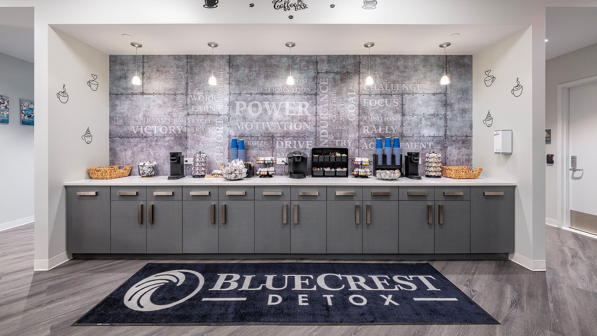 A coffee and refreshment station at BluffCrest Detox featuring a gray counter, kitchen appliances, and a branded floor mat.