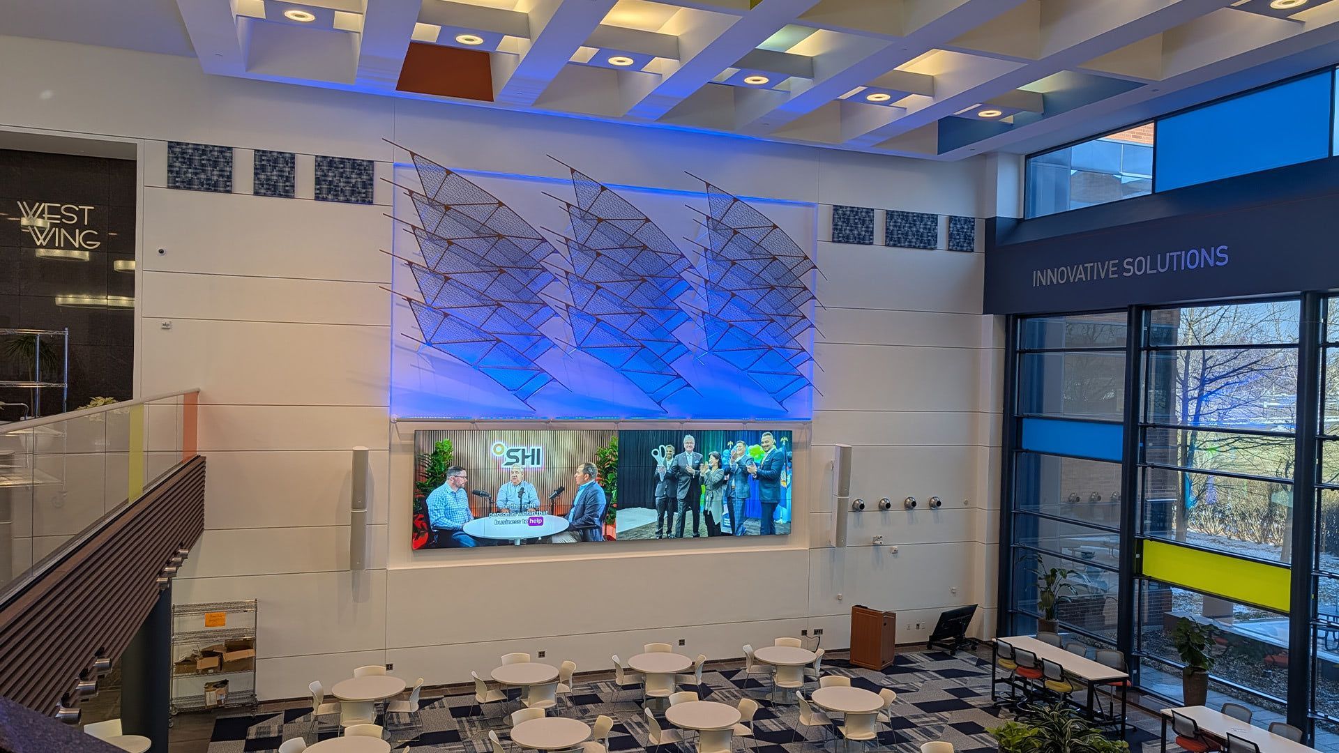 A spacious, modern atrium features a large central screen displaying a video, accented by blue lighting and white seating.