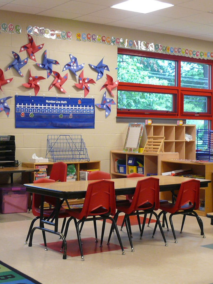 A classroom scene with a table surrounded by red chairs, beneath a wall decorated with red and blue paper pinwheels.