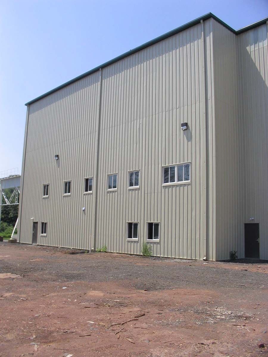 A side view of a tall, beige, corrugated metal industrial building with several small windows under a clear blue sky.