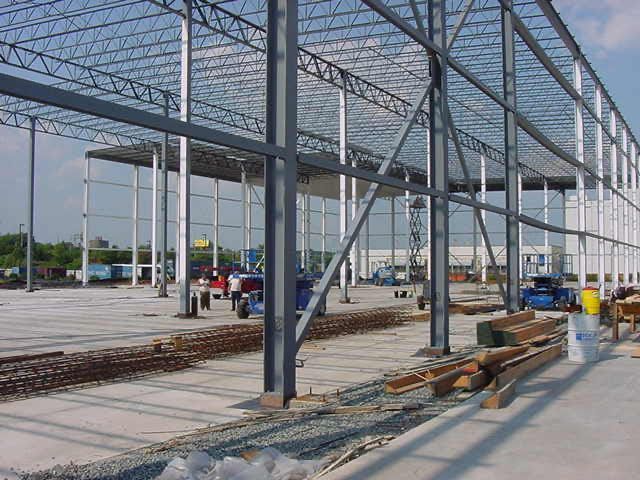 An outdoor construction site showing the steel framework of a large industrial warehouse under a clear blue sky.