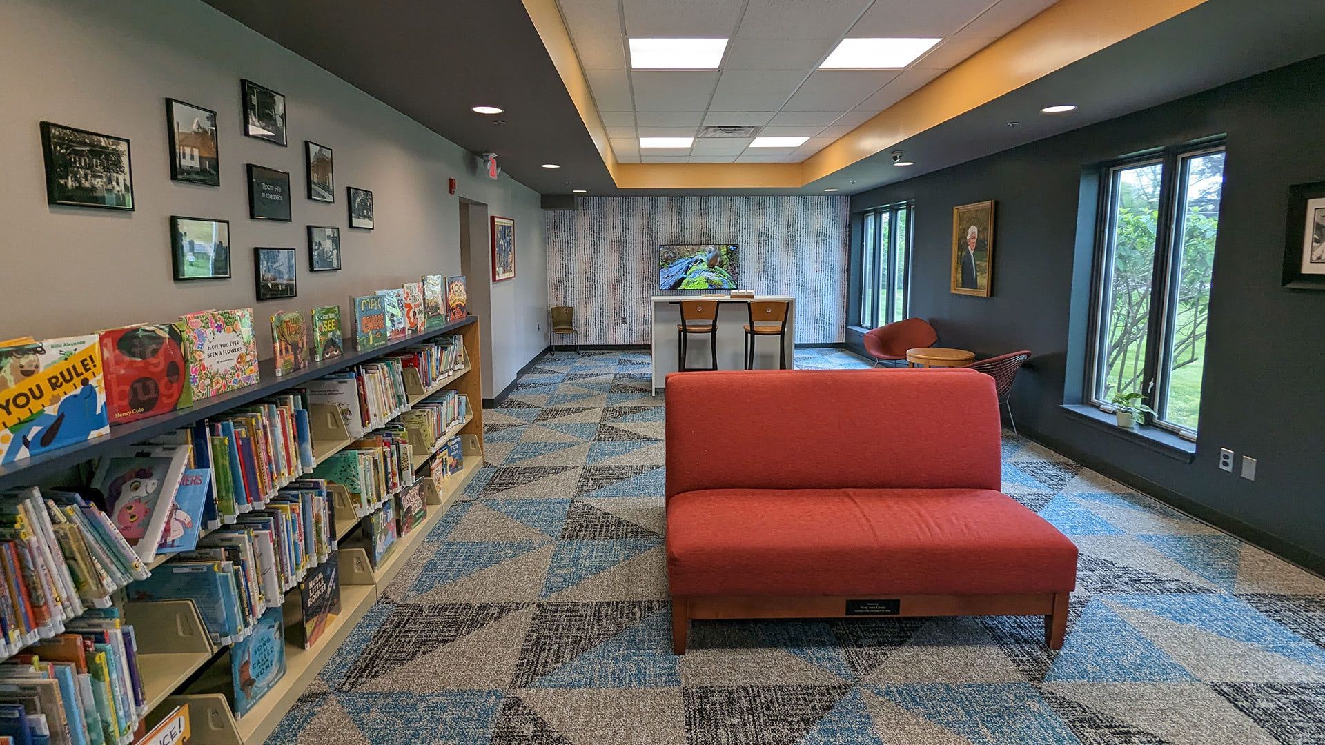 A colorful library area with rows of books on shelves to the left, a bright red sofa in the center, and patterned carpet.