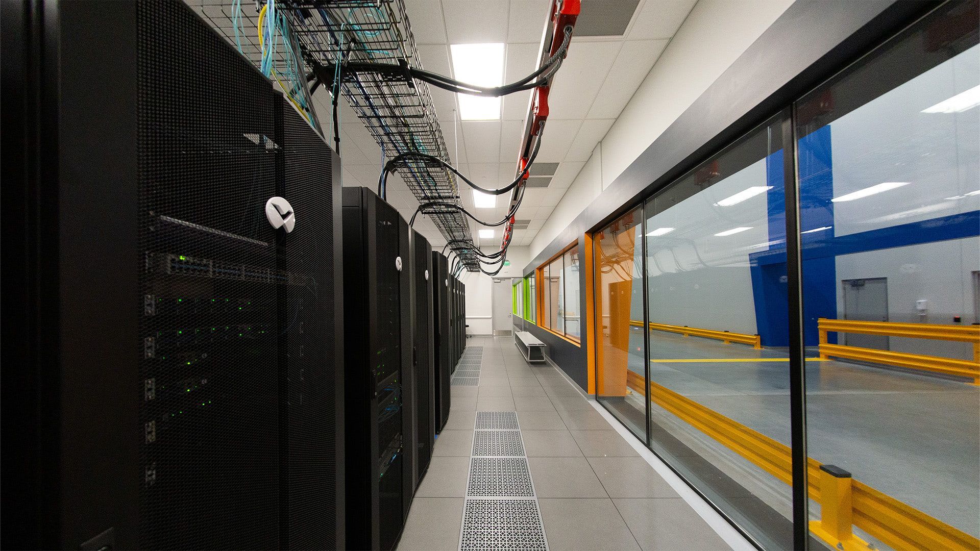 A row of black server cabinets lines a hallway next to a glass wall overlooking a large, industrial facility.