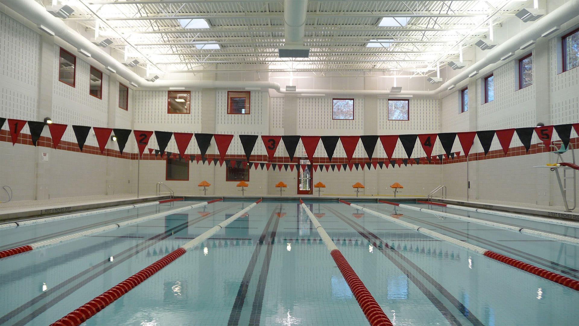 An empty indoor swimming pool with multiple lanes, red and black pennant banners, and white walls with small windows.