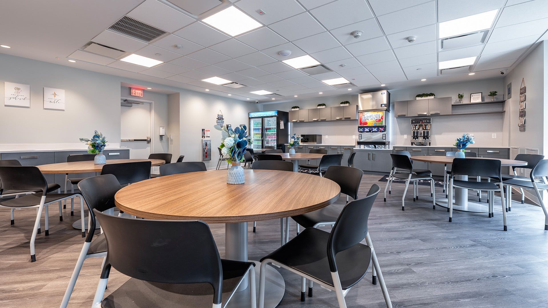 A bright office breakroom featuring multiple round wood-topped tables surrounded by black chairs and a kitchen area.