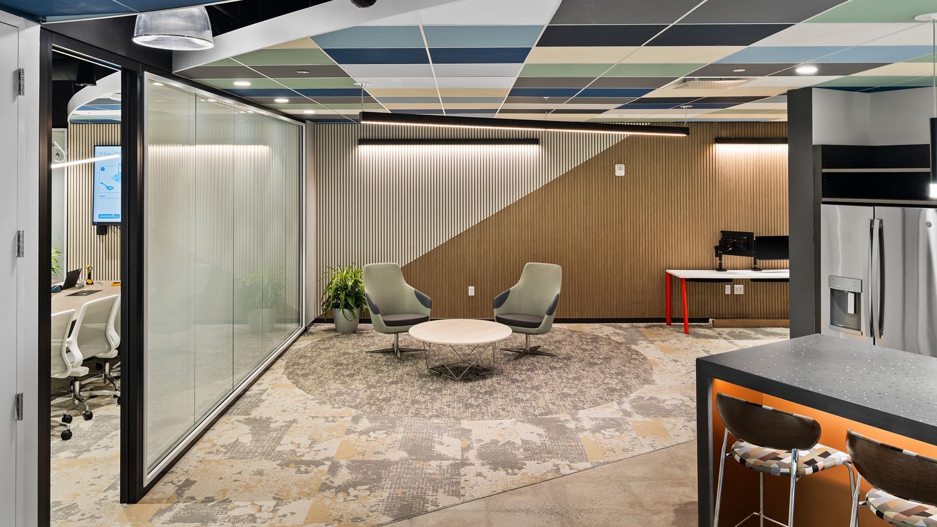 A modern office lounge featuring two armchairs, a round coffee table, a glass-walled conference room, and a kitchenette.