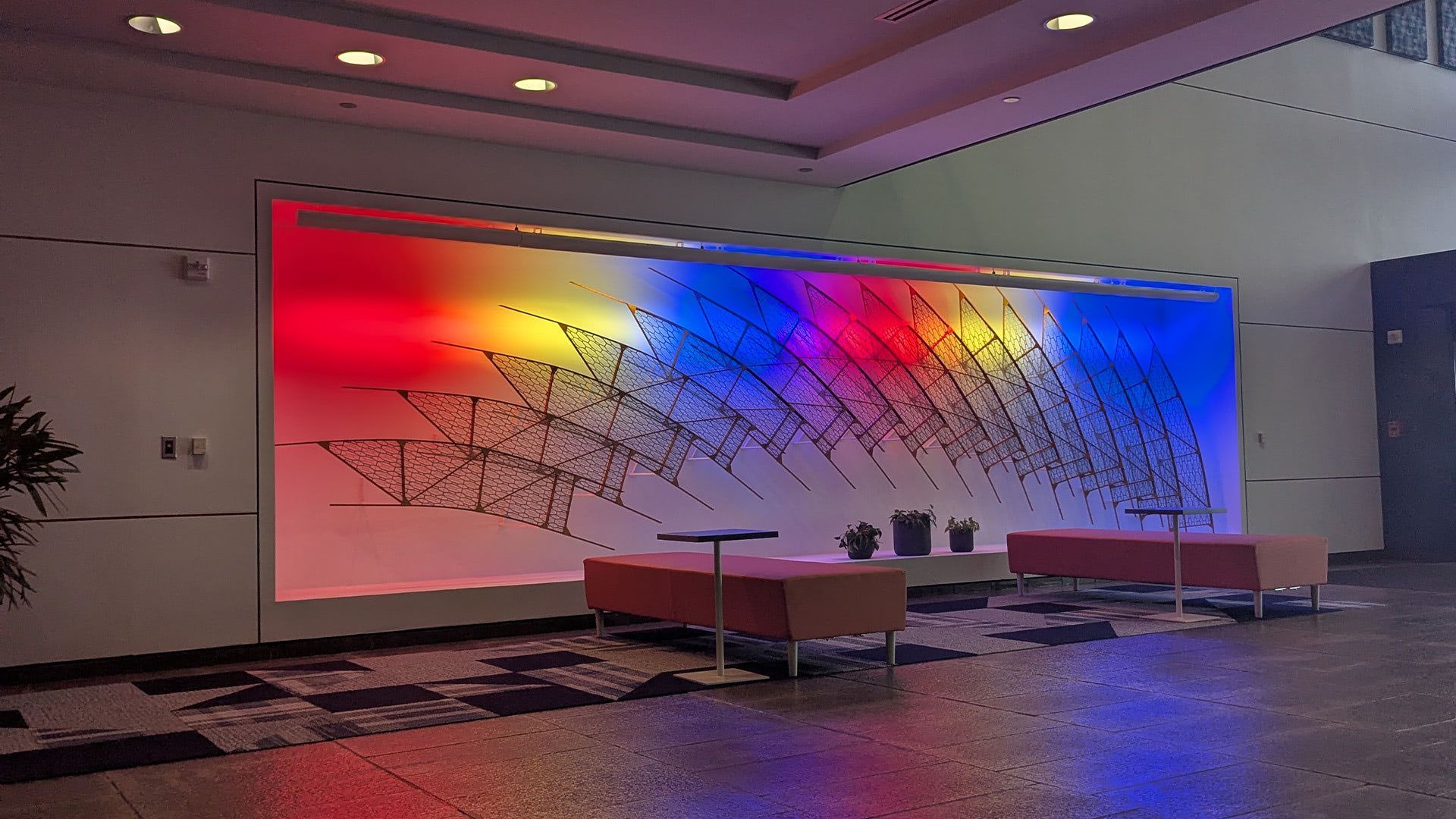 A lobby featuring a large wall mural with gradient rainbow lighting behind an abstract, dark metal wire structure.