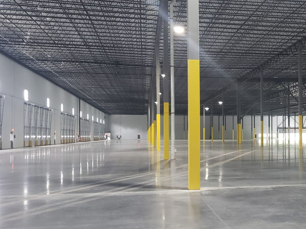 A vast, empty warehouse with polished concrete floors, high ceilings, and rows of columns with yellow-painted bases.